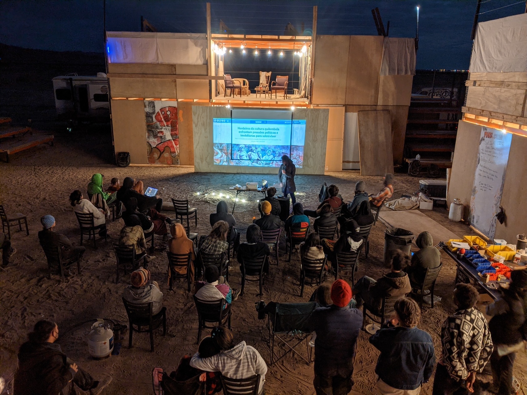 An outdoor presentation at night with a crowd watching a projector screen.