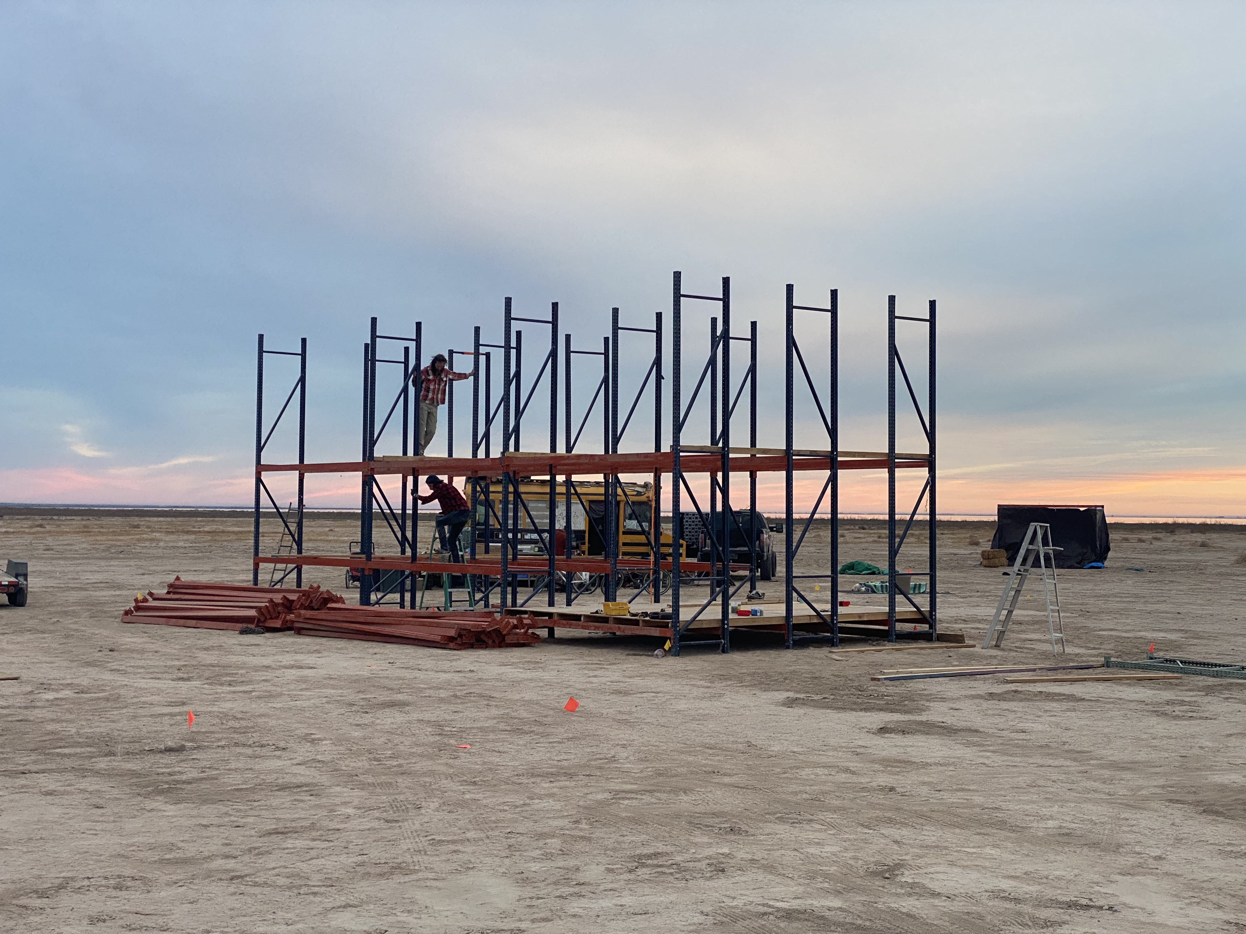 Two people constructing a large pallet rack structure in the desert at dusk.