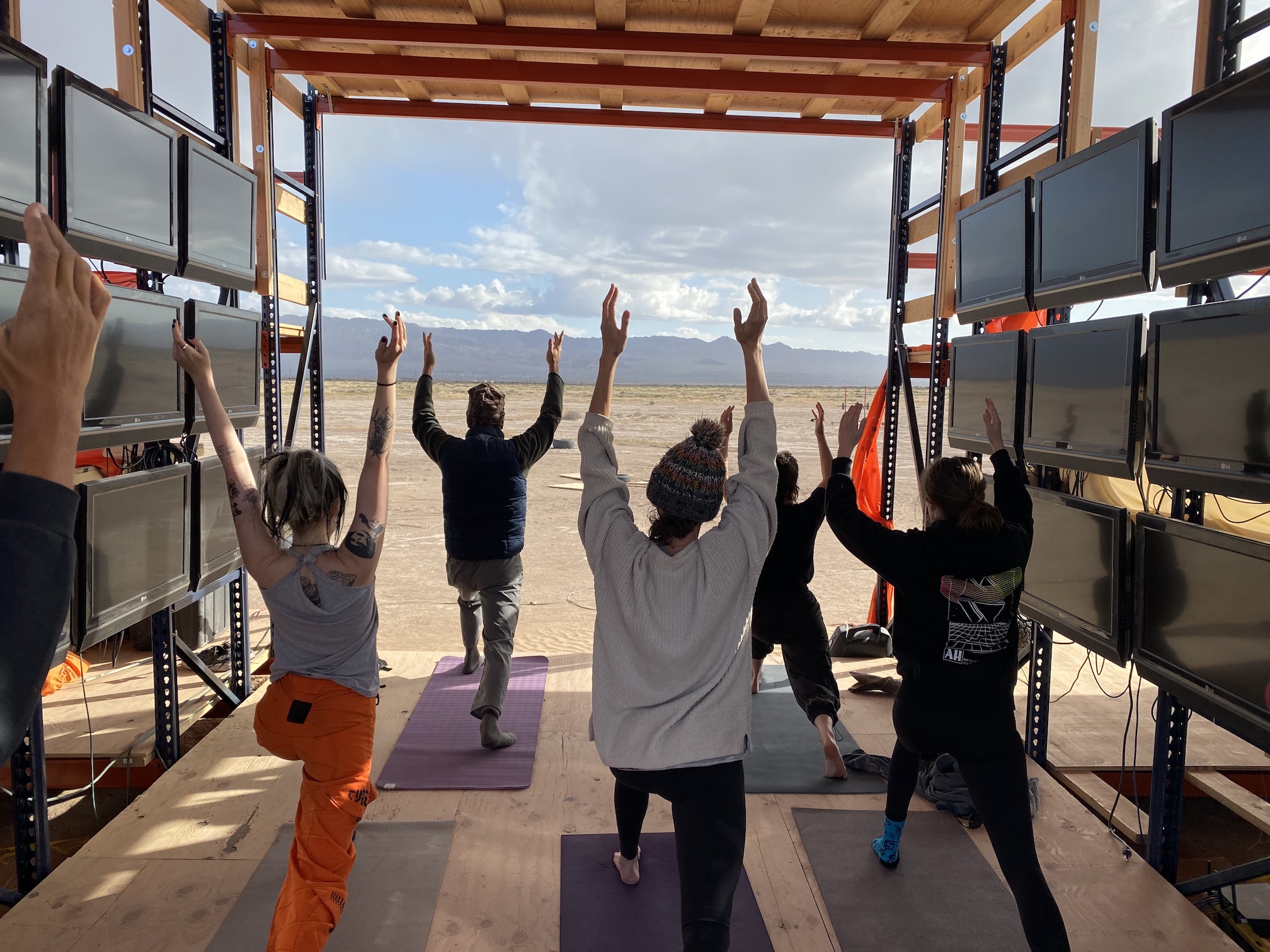 A group does yoga inside a pallet-rack structure, looking out at the desert.