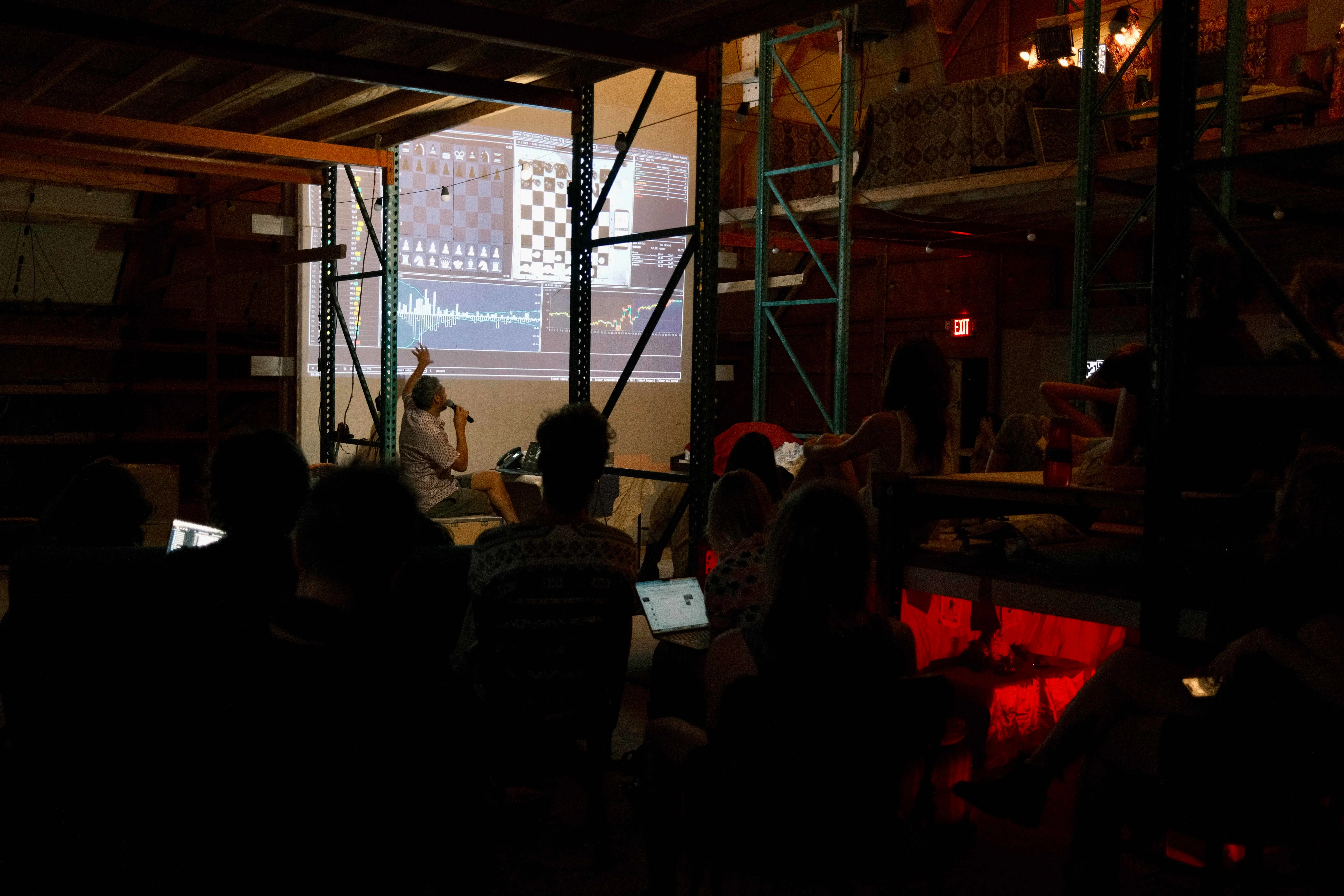 A Thunder Talk in the Chiba hangar at night — a presenter with a microphone in front of a projection of chess engines and live charts, audience watching from the dark