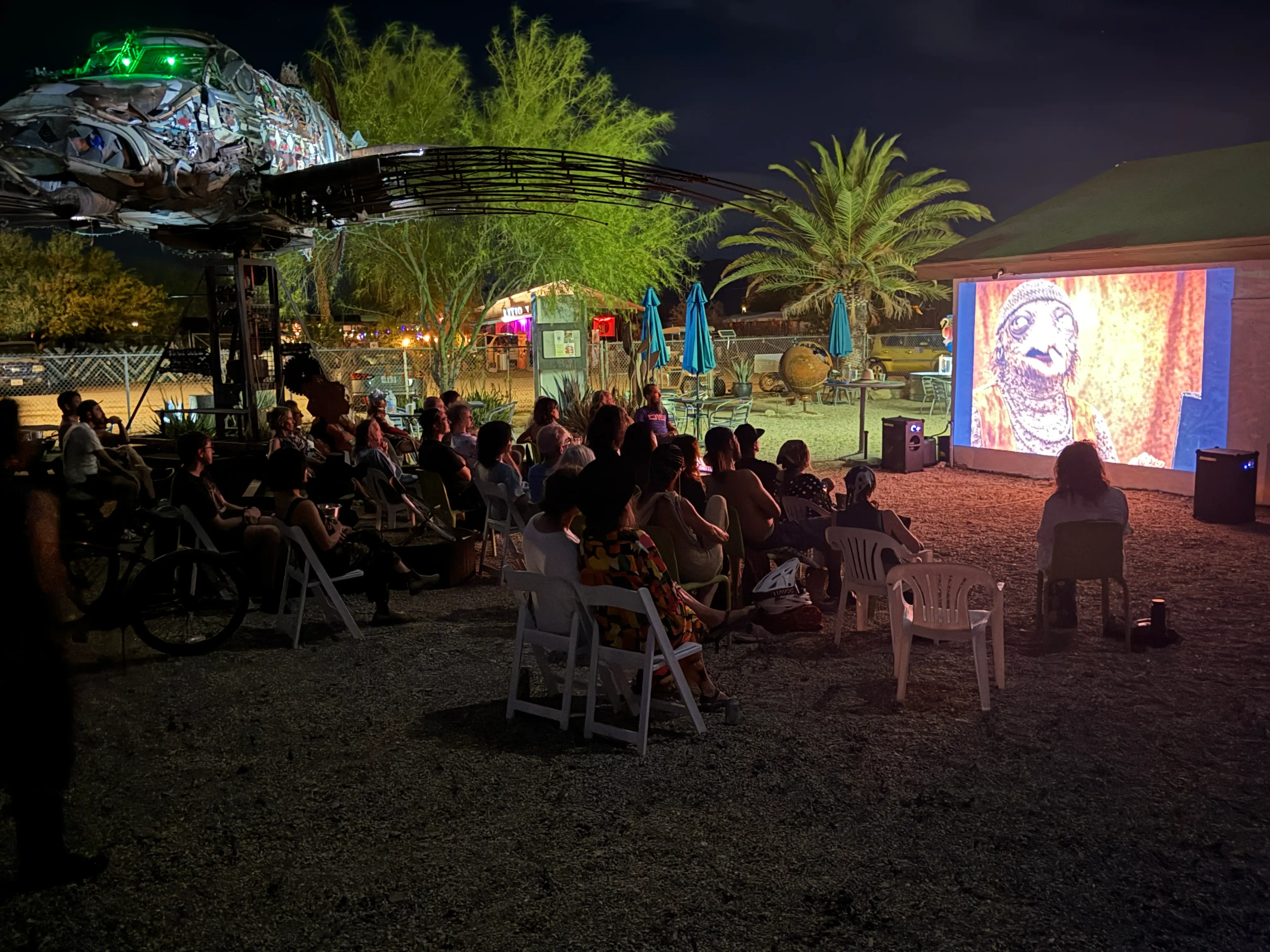 The 2026 AI Film Festival screened outdoors at the Bombay Beach Arts Center — audience in folding chairs under the recycled-metal fish sculpture, palm tree beside the glowing screen