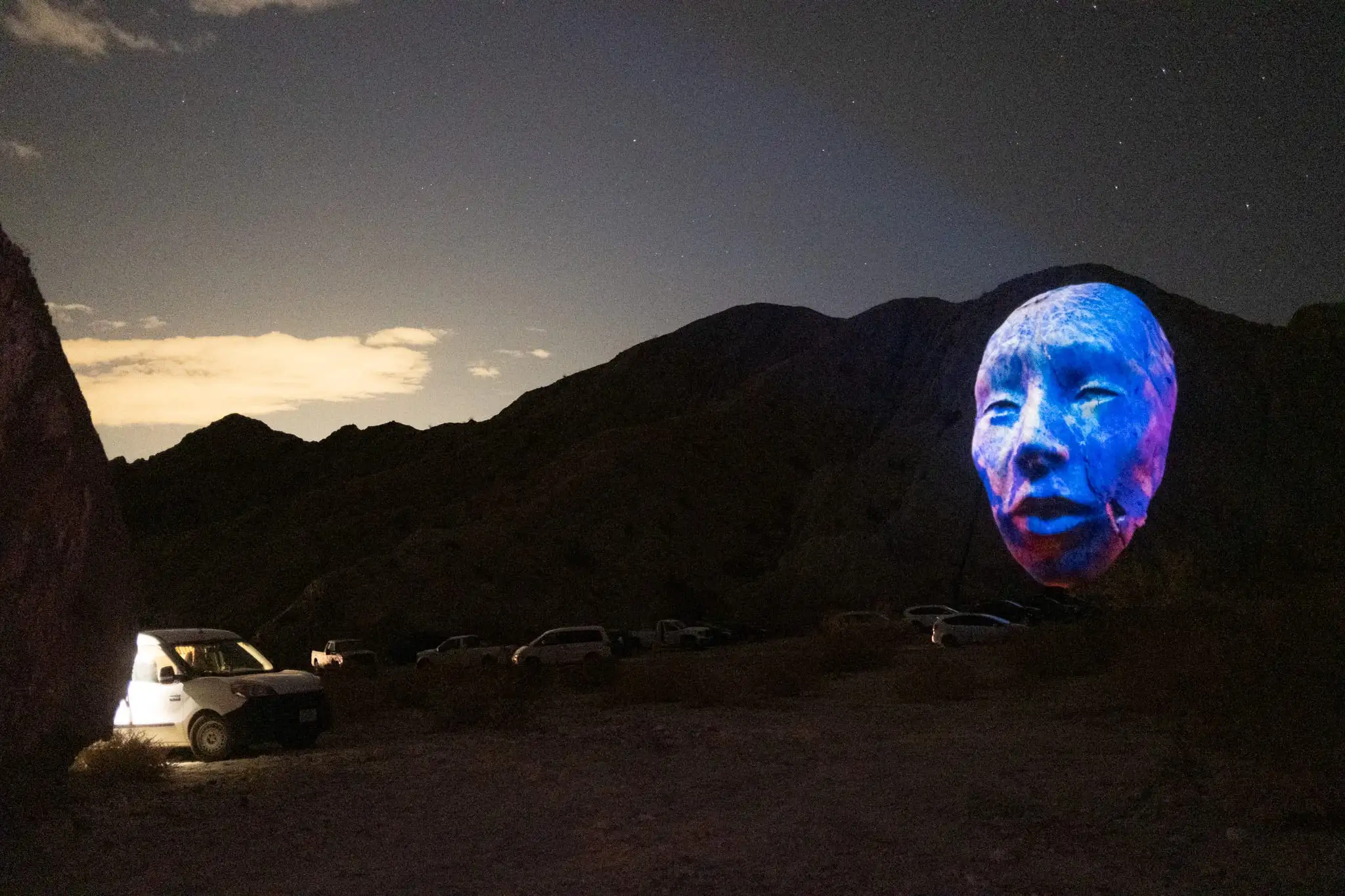 A giant AI-generated face projected onto a canyon wall at night, cars parked on the desert floor below — Painted Canyons, 2025