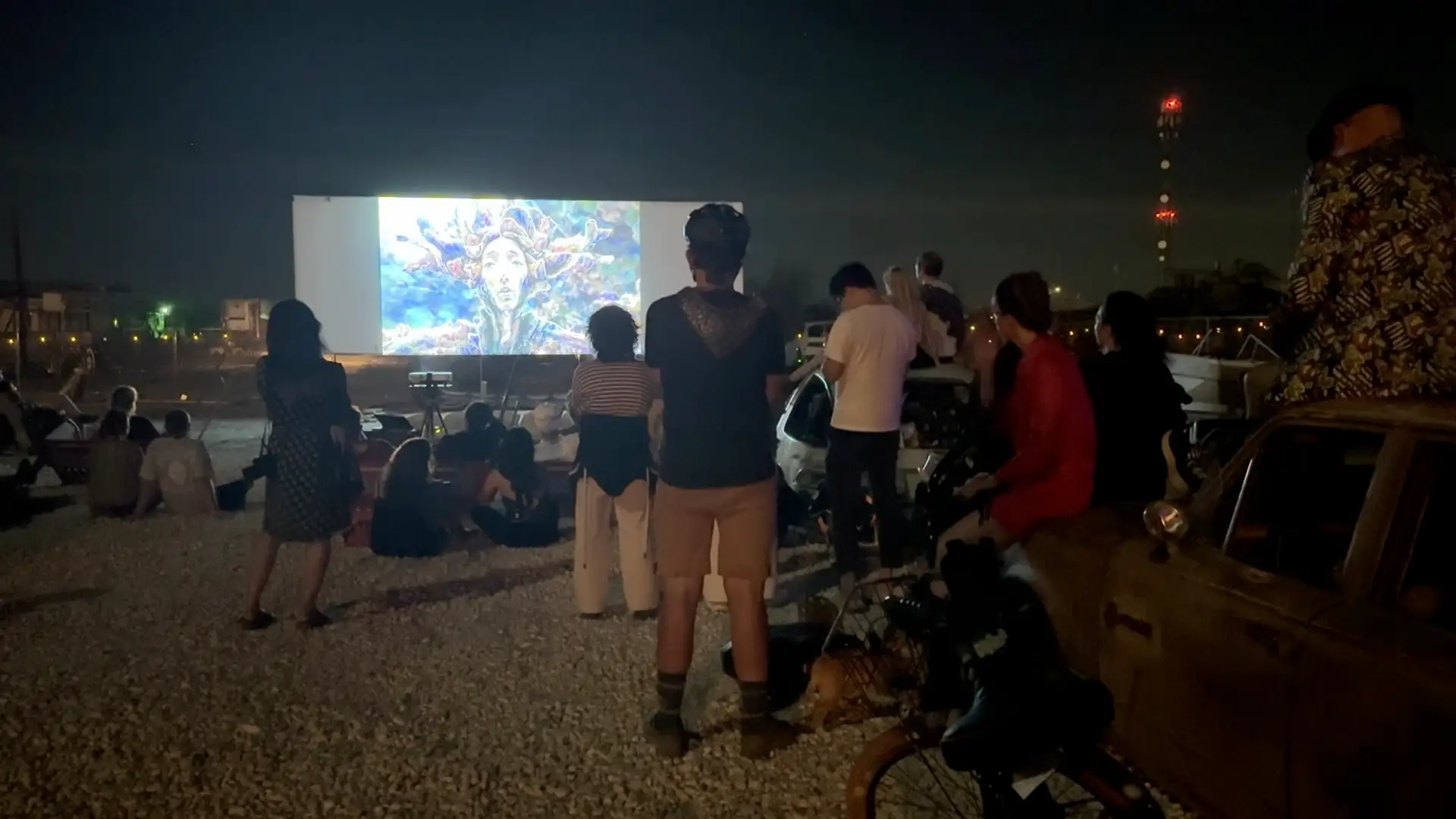 A standing crowd watching a generative AI film at the drive-in, radio tower blinking behind · 2025