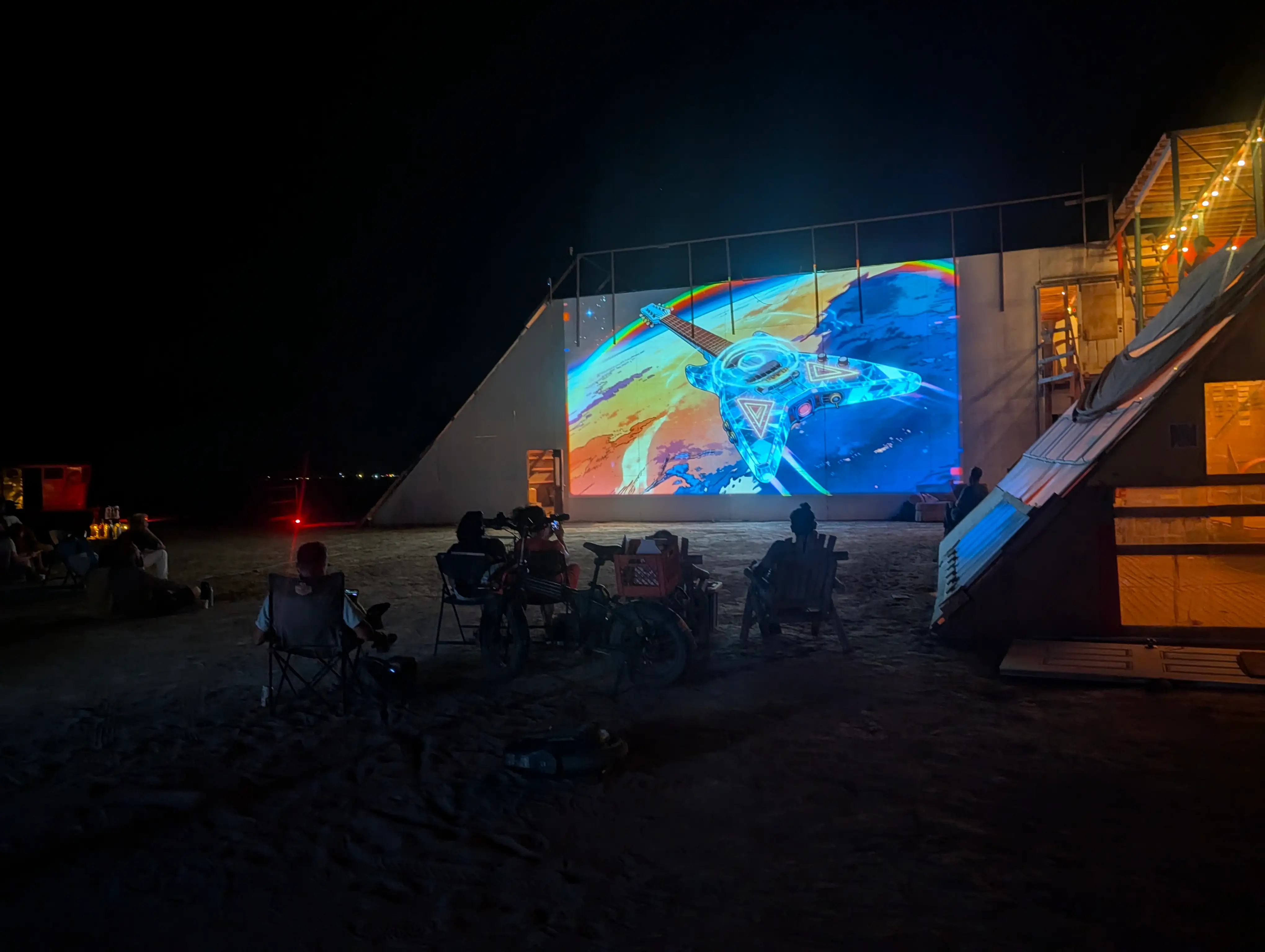 A glowing guitar-shaped spaceship projected across a pyramid wall at Mars, the audience watching from camp chairs · 2026