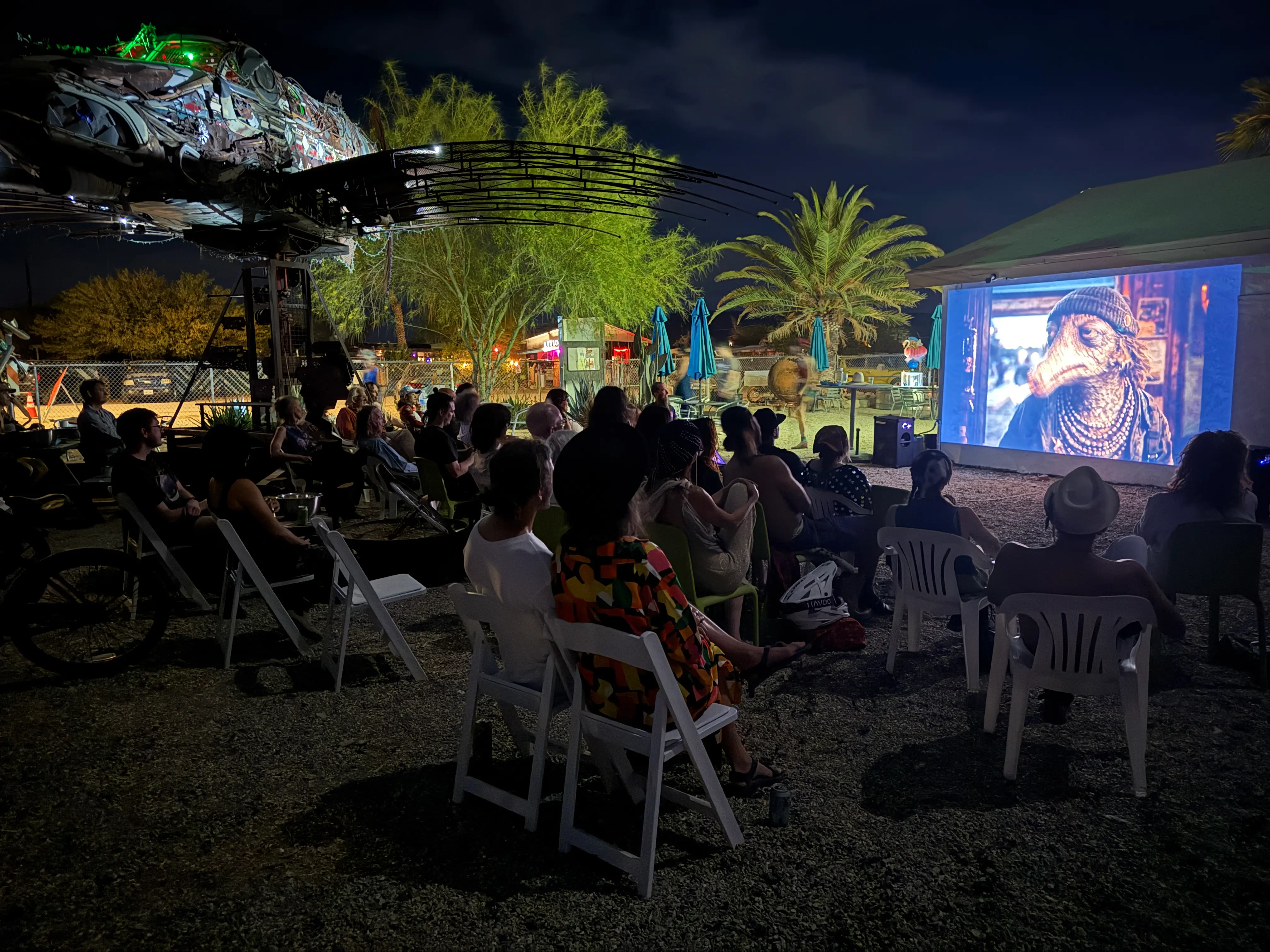 An outdoor night screening at the Bombay Beach Arts Center — audience in folding chairs, palm tree beside the glowing screen, recycled-metal fish sculpture overhead