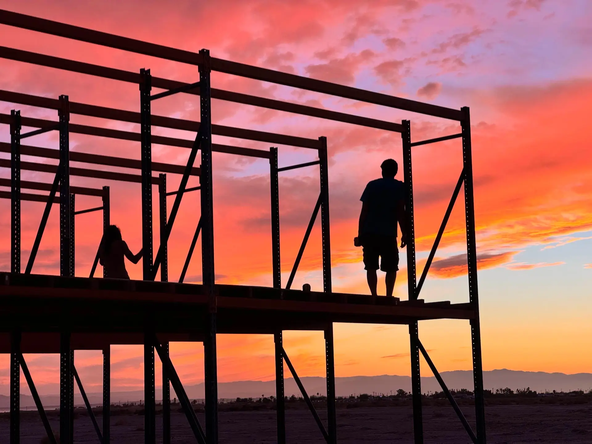 Two people silhouetted on a pallet-rack structure against a vivid desert sunset